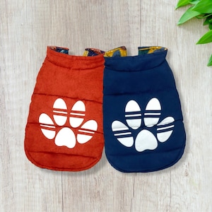 May include: Two dog coats, one red and one blue, with a white paw print design on each. The coats are quilted and have a zipper closure.