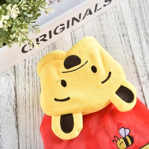 May include: Red hooded plush toy with a yellow Winnie the Pooh head and brown ears. The hood has a smiling face with black eyes and a black nose. The toy has a small embroidered bee on the front.