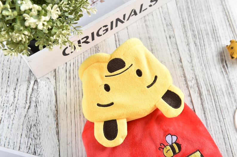 May include: Red hooded plush toy with a yellow Winnie the Pooh head and brown ears. The hood has a smiling face with black eyes and a black nose. The toy has a small embroidered bee on the front.
