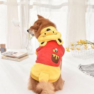 May include: A red and yellow dog costume with a Winnie the Pooh design. The costume features a hood with bear ears and a bee and honey pot design on the back.