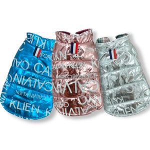 May include: Three puffer jackets for dogs in blue, pink, and silver with the words "Calvin Klein" repeated on the fabric.