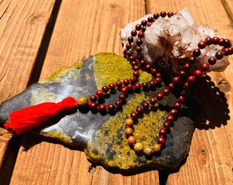 May include: A mala bead necklace with reddish-brown beads and a red tassel. The necklace rests on a multi-colored stone with a clear quartz crystal in the background. The beads are strung together, creating a long, elegant strand.