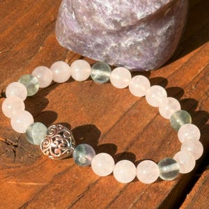 Puede incluir: Un brazalete hecho con cuentas rosas y verdes con un dije de corazón plateado.