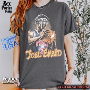 May include: Dark gray t-shirt featuring a graphic of a masked figure holding a basketball, with "PHIL" and "JOEL EMBID" text. The shirt is labeled "Comfort Colors 1717" and is available in oversized sizes.