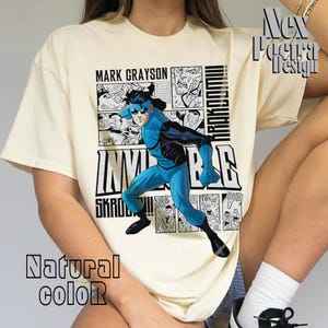 Puede incluir: Una camiseta de color crema con un gráfico de cómic de un superhéroe azul con el texto "Mark Grayson" e "Invincible" impreso en la parte delantera.