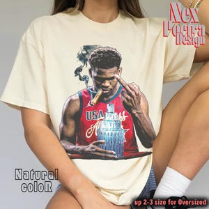 Pode incluir: Camiseta creme com um gráfico de um jogador de basquete fumando um charuto e mostrando o dedo do meio. O jogador está vestindo uma camisa dos EUA vermelha e azul. A camisa tem o texto "Natural color" e "up 2-3 size for Oversized".