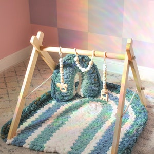 May include: A wooden A-frame baby play gym featuring a thick, crocheted, blue and white striped mat and a matching arch-shaped pillow. Three hanging toys made of wooden beads and small wooden animal shapes are suspended from the top bar.
