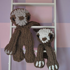 May include: Two brown and white knitted sloth toys on a white shelf. The larger sloth is hanging from the shelf with its arms outstretched. The smaller sloth is sitting on the shelf with its legs dangling.