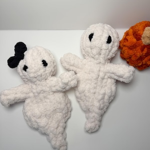May include: Two white crocheted ghost plushies made of soft, chunky yarn. One ghost features a small black bow on its head, while both have simple black embroidered eyes. A portion of a textured orange crocheted pumpkin is visible on the right side.