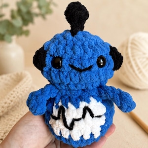 Può includere: Un piccolo peluche rotondo lavorato all'uncinetto di colore blu, con un ciuffo nero sulla sommità, orecchie nere e due occhi neri. Il corpo presenta un motivo bianco a denti di sega al centro con una linea nera a zigzag. Il testo Melinda Murrin è visibile.