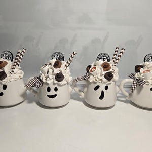Op de afbeelding: Vier witte keramische mokken met spookgezichten, gegarneerd met slagroom, snoep en rietjes. Elke mok heeft een oor en een zwart-wit geblokte strik. Bovenop elke mok staat een bordje met "FRESHLY BREWED COFFEE".