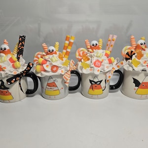 May include: Four Halloween-themed mugs with black handles and rims. Each mug features a candy corn character design and is topped with whipped cream, candies, and decorative straws. The mugs are decorated with a witch hat, a party mask, and a bat wing.