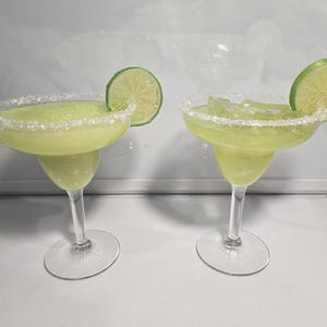 Könnte beinhalten: Zwei grüne Margarita-Cocktails mit einem gesalzenen Rand und Limettenspalten. Die Cocktails befinden sich in klaren Glas-Margarita-Gläsern auf einer weißen Oberfläche.