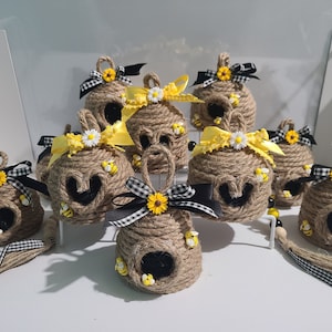 Könnte beinhalten: Handgefertigte dekorative Bienenkorb-Ornamente aus Bindfaden, verziert mit gelben Bändern, Gänseblümchen und Miniatur-Bienen. Jedes Ornament hat eine schwarz-weiß karierte Schleife und eine Quaste aus Jute. Perfekt für die Frühlings- oder Sommerdekoration.