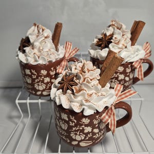 May include: Three decorative mugs filled with whipped cream, cinnamon, and star anise. Each brown mug features a floral and pumpkin design and is adorned with a cinnamon stick and a red and white checkered ribbon. The mugs are displayed on a white wire shelf.