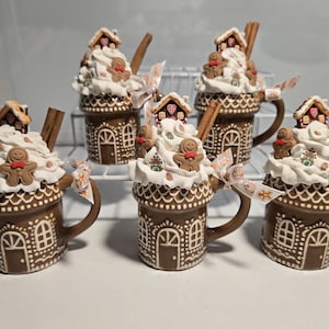 May include: A collection of six brown ceramic mugs designed as gingerbread houses, topped with white frosting, gingerbread men, and miniature houses. Each mug has a cinnamon stick and is adorned with a decorative ribbon.