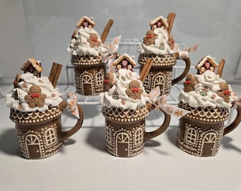 May include: A collection of six brown ceramic mugs designed as gingerbread houses, topped with white frosting, gingerbread men, and miniature houses. Each mug has a cinnamon stick and is adorned with a decorative ribbon.