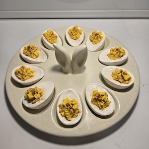 May include: A round, speckled off-white ceramic deviled egg serving platter featuring a central handle shaped like two upright rabbit ears. The platter holds ten individual egg-shaped indentations, each containing a halved egg with yellow filling.