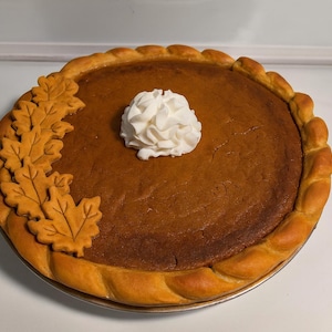 Op de afbeelding: Een hele pompoentaart met een goudbruine korst en vulling. De taart is versierd met een cluster van bladvormige gebakken uitsteeksels en een toef slagroom in het midden. De taart staat op een zilveren taartbord.