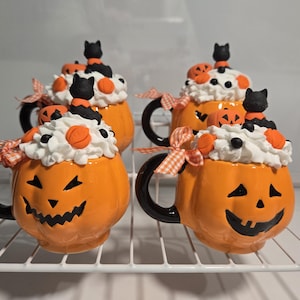 May include: Four orange pumpkin-shaped mugs with black handles. Each mug is decorated with white frosting, orange and black candy, and a black cat figurine.