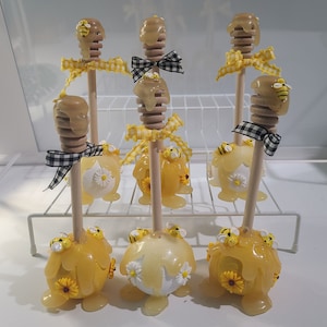 Puede incluir: Seis palitos de miel decorados con abejas amarillas y blancas, flores amarillas y blancas y una cinta a cuadros negros y blancos. Los palitos están dispuestos en una rejilla de alambre blanca.