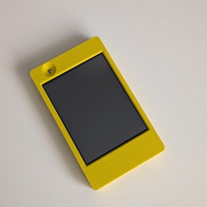 May include: A rectangular electronic device featuring a bright yellow textured frame surrounding a dark, blank screen. The frame includes a circular cutout in the upper left corner, revealing a small metallic component beneath the surface.