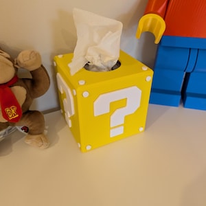 May include: A yellow tissue box shaped like a question mark block from the Super Mario Bros. video game series. The box has white question marks and white dots on the corners. White tissues are coming out of the top.
