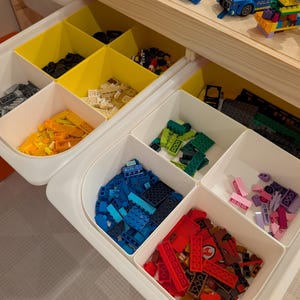 May include: A white storage unit with yellow and white compartments filled with colorful toy building blocks. The blocks are in various colors, including blue, red, green, yellow, and pink. A wooden shelf above holds assembled toy cars.