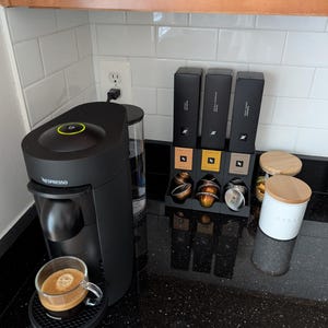 May include: A black Nespresso Vertuo coffee machine with a glass of espresso on a black coaster. Three black boxes of coffee pods and a white container with a wooden lid are on the countertop. The countertop is black with white flecks.