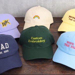 May include: Six baseball caps in different colours, including navy, lavender, and yellow. Each cap has embroidered text or designs, such as "DAD", a rainbow, and "WIFE OF THE PARTY".