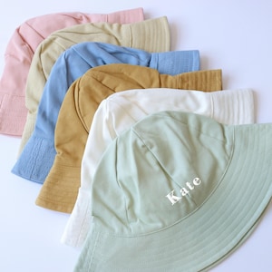 May include: A stack of six colourful bucket hats, with the top hat being a light green colour and the text "Kate" embroidered on the front. The hats are made of a soft, cotton fabric.