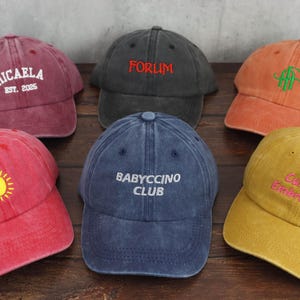 May include: Six baseball caps in various colours, including red, orange, blue, and yellow, with embroidered text and designs. The caps feature text such as "Micaela", "Forum", and "Babyccino Club".