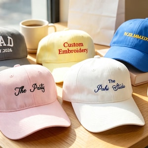 May include: Five baseball caps in grey, yellow, blue, pink, and white. The grey cap features white text reading DAD EST.2026. The yellow cap has red text reading Custom Embroidery. The blue cap has yellow text reading IV.XII.MMXIX. The pink cap has black text reading The Party, and the white cap has blue text reading The Pahi Studio.