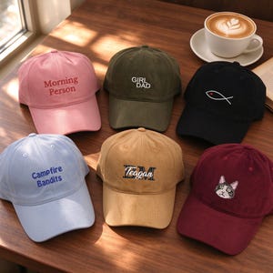 May include: Six baseball caps in various colours including pink, olive green, black, light blue, tan, and maroon. The caps feature embroidered designs: Morning Person, GIRL DAD, a fish symbol, Campfire Bandits, the name Teagan, and a cat face.