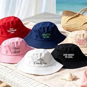 May include: A collection of six bucket hats in red, navy blue, light pink, beige, white, and black. Each hat features custom embroidery, including text such as Your Text/Design here, SMITH Est. 1892, Team Bride, Anna's Bachelor Bash, and LIVE MUSIC IS COOL.