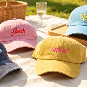 May include: Four baseball caps in navy blue, pink, mustard yellow, and light blue. The navy cap features the name Diana in multicoloured embroidery. The pink cap has the name Josch in red. The mustard cap says Minki in pink, and the light blue cap displays the yellow monogram TL.