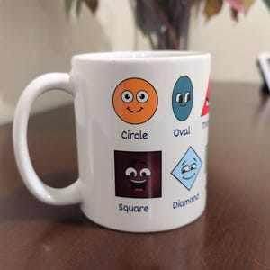 Pode incluir: Caneca de cerâmica branca com um gráfico colorido de diferentes formas com rostos. As formas incluem um círculo, um oval, um quadrado, um diamante, um triângulo e uma estrela. Cada forma tem uma cor diferente e um rosto sorridente. O texto abaixo de cada forma identifica a forma. A caneca está sobre uma superfície de madeira marrom.