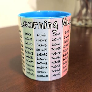 Puede incluir: Una taza de cerámica azul y blanca con las palabras "Learning Mug" impresas en la parte delantera. La taza tiene un diseño colorido con tablas de multiplicar impresas en ella. Las tablas incluyen 5x1 a 5x10, 6x1 a 6x10, 7x1 a 7x10 y 8x1 a 8x10.