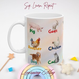 Op de afbeelding: Een witte keramische mok met een kleurrijk ontwerp met verschillende dieren en hun namen, waaronder een varken, een geit, een haan, een kip, een hond en een kat. De mok heeft de tekst "Sip, Learn, Repeat" en "Learning Mug" erop gedrukt.