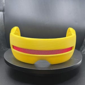 May include: A bright yellow visor with a horizontal cutout across the center. The cutout features a textured, metallic red insert with a fine diamond-patterned grid. The visor has a curved, wrap-around shape with smooth, solid yellow edges.