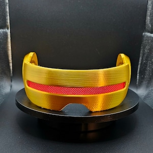 May include: A gold-colored visor with a red, textured stripe across the front. The visor is on a black circular stand.