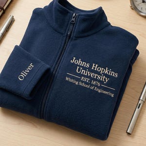 Puede incluir: Chaqueta polar azul marino con cremallera, con el texto "Johns Hopkins University" y "Oliver" bordado en blanco. También se ven un bolígrafo plateado, un cuaderno y un reloj pequeño.