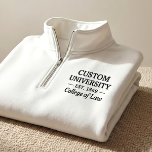 Könnte beinhalten: Ein gefaltetes weißes Sweatshirt mit einem Kragen mit Viertelreißverschluss. Das Sweatshirt trägt den schwarzen Text "CUSTOM UNIVERSITY - EST. 1869 - College of Law". Der Reißverschluss ist silberfarben.