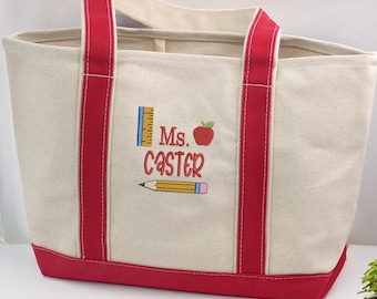 May include: A canvas tote bag with red accents. The bag features red handles and a red base. The front of the bag has a colorful graphic with a ruler, apple, pencil, and the text "Ms. Caster".