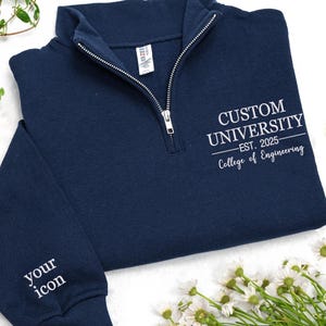 Peut inclure: Sweat-shirt bleu marine à fermeture éclair quart de zip avec texte brodé blanc. Le texte sur le devant indique "CUSTOM UNIVERSITY - EST. 2025 - College of Engineering." La manche porte le texte "your icon." La fermeture éclair est argentée.