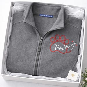 May include: A gray fleece zip-up jacket with a red paw print and stethoscope design, embroidered with the name "Terri". The jacket is presented in a white gift box with tissue paper.
