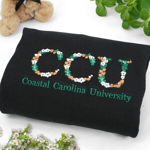 Może przedstawiać: Czarny koc z haftem "CCU" w kwiatowym wzorze z bieli, zieleni i brązu. Słowa "Coastal Carolina University" są wyhaftowane poniżej w kolorze zielonym.