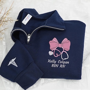 May include: Navy blue quarter-zip sweatshirt with a silver zip. The sweatshirt features a pink bow and stethoscope design, along with the name "Kelly Colgan BSN RN" embroidered in white. A caduceus is embroidered on the left sleeve.