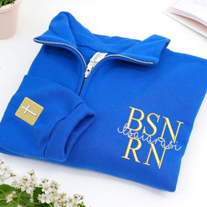 May include: Royal blue zip-up sweatshirt with gold embroidered text reading "BSN RN" and "Tsaiarasi". A gold square patch with a white cross is on the left sleeve. The sweatshirt has a high collar and a silver zipper.