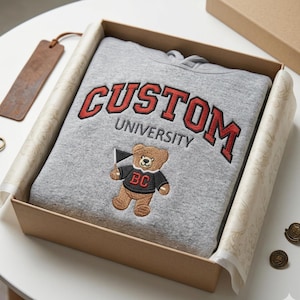 A Gildan Sport Grey hoodie neatly folded inside a luxury kraft gift box with premium tissue paper. The custom university embroidery features red "CUSTOM" lettering and an embroidered bear mascot. Ideal for personalized graduation gifts or alumni homecoming presents, this high-end presentation highlights the quality of custom-made collegiate outerwear.